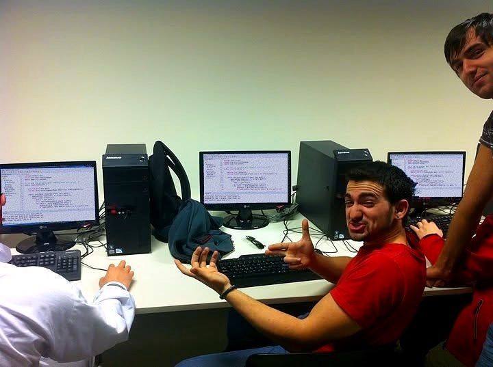 Une salle informatique à l’université : Nikos se tourne vers l’objectif devant une rangée d’écrans affichant du code.